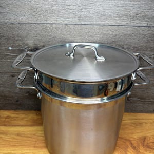 Könnte beinhalten: Edelstahl-Kochtopf mit Deckel und Dampfeinsatz. Der Topf hat zwei Seitengriffe und einen Griff am Deckel. Der Dampfeinsatz sitzt im Topf. Der Topf steht auf einer Holzoberfläche.