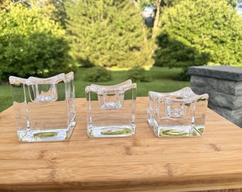 May include: Three clear glass candle holders of varying heights, each featuring a square base and a concave top surface with a central circular opening designed to hold a taper candle securely in place within the thick, transparent glass structure.
