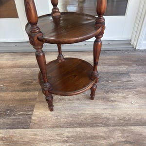 Könnte beinhalten: Ein zweistöckiger, dunkelbrauner Pflanzenständer aus Holz mit glänzendem Finish. Der Ständer verfügt über zwei kreisförmige Regale, die von gedrechselten Beinen und einer Mittelsäule getragen werden. Der Ständer ist etwa 76 cm hoch.