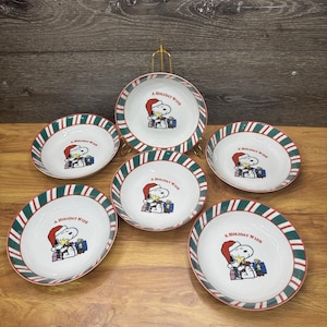 May include: Set of six white ceramic bowls with a red and green striped border. Each bowl features a Snoopy graphic wearing a Santa hat, holding gifts, and the text "A Holiday Wish".