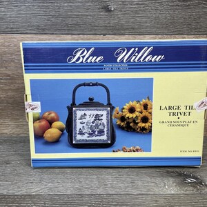May include: A Blue Willow pantry collection trivet in a box. The trivet has a black frame and a blue and white design. The box also features a bouquet of sunflowers and some fruit.