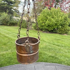 May include: A cylindrical copper hanging planter featuring a weathered patina and a horizontal ridge around the center. The vessel is suspended by three rusted metal chains attached to decorative brass-toned triangular brackets.