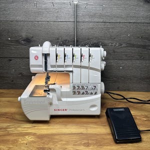 Puede incluir: Una remalladora Singer Professional 5 blanca con soporte de hilo de metal, cinco diales de tensión y luz integrada. La máquina presenta una guía de puntadas impresa y el texto Singer Professional 5. Se incluye un pedal electrónico de color negro.