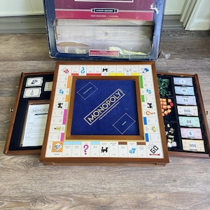 Op de afbeelding: Een Monopoly Luxury Edition bordspelset met een houten kast met een blauw vilten middenstuk, een speelbord met eigendomsvakken, metalen pionnen, papieren speelgeld en plastic huizen en hotels. De set bevat een grote kartonnen doos.