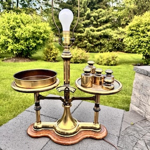 May include: Vintage brass balance scale converted into a lamp. The lamp has a large light bulb, a round tray, and a set of brass weights. The base is made of wood and the lamp is set on a stone surface.