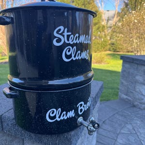 Op de afbeelding: Zwarte, gespikkelde emaille stoompan voor mosselen met de woorden "Steamed Clams" en "Clam Broth" in witte letters. De pan heeft een kraan en een deksel. De achtergrond is een grasveld met bomen.