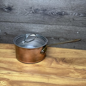 May include: A copper-colored saucepan with a long, brass-colored handle and a clear lid. The pot sits on a wooden surface with a wood grain pattern. The background is a gray wooden wall.