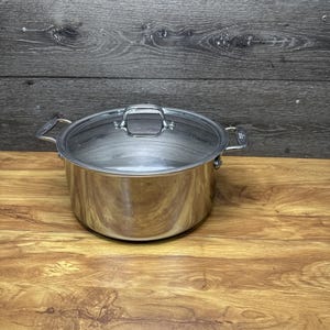 Könnte beinhalten: Ein Edelstahltopf mit Glasdeckel. Der Topf hat zwei seitliche Griffe und einen Griff am Deckel. Der Topf steht auf einer Holzoberfläche.