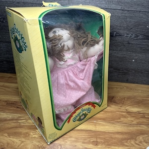May include: A vintage Cabbage Patch Kids doll in its original box. The doll has brown yarn hair and is wearing a pink dress with white polka dots and lace trim. The box is yellow with green accents and the Cabbage Patch Kids logo.