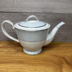 Może przedstawiać: Biały i jasnoniebieski porcelanowy imbryk z pokrywką, uchwytem i dzióbkiem. Imbryk ma srebrne wykończenie i kwiatowy wzór. Imbryk stoi na drewnianej powierzchni.