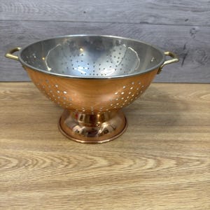 May include: A copper and stainless steel colander with a pedestal base and two brass handles. The colander has a bowl shape with small holes for draining liquids. The copper exterior contrasts with the stainless steel interior.
