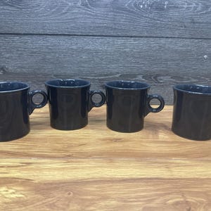 Op de afbeelding: Vier marineblauwe keramische mokken met afgeronde handvatten staan op een houten oppervlak. De mokken hebben een eenvoudig, klassiek ontwerp, met een licht uitlopende rand. De mokken zijn gelijkmatig verdeeld.