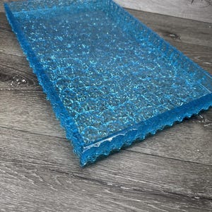 May include: A rectangular, transparent blue tray with a textured surface and a scalloped edge. The tray is made of a clear material, and the surface has a bubbly, irregular pattern. The tray is displayed on a wooden surface.