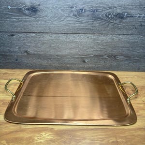 May include: A rectangular copper serving tray with rounded corners and two curved brass handles attached to the sides. The tray features a polished, reflective metallic surface with a warm, reddish-brown tone and smooth, clean edges throughout.