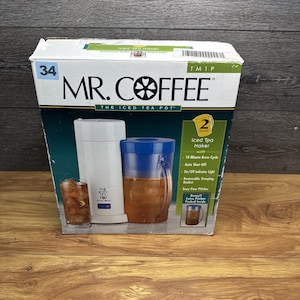 以下が含まれることがあります： Mr. Coffee のアイスティーメーカーの箱。箱にはブランド名と製品の詳細が表示されており、容量は1.9リットルです。画像には、ティーメーカー、ピッチャー、アイスティーのグラスが写っています。