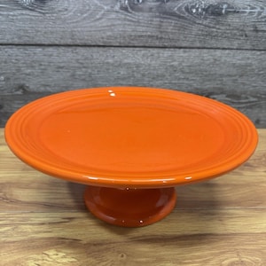 May include: A bright orange ceramic cake stand featuring a smooth, glossy finish with a wide, circular top plate and a sturdy, pedestal-style base. The design includes subtle concentric ring detailing along the outer edge of the plate surface.