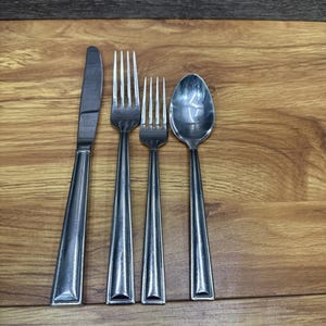 Könnte beinhalten: Ein Besteckset aus Edelstahl, bestehend aus einem Messer, zwei Gabeln und einem Löffel. Das Besteck liegt auf einer Holzoberfläche. Das Messer befindet sich links, gefolgt von einer großen Gabel, einer kleineren Gabel und einem Löffel.