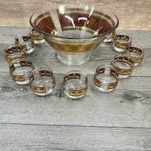 May include: A vintage glass punch bowl set with a large bowl and twelve small glasses. The set features gold and brown decorative accents. The glasses are arranged around the bowl on a wooden surface.