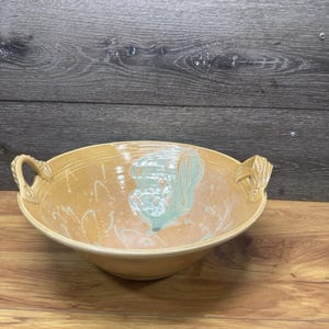 Puede incluir: Un cuenco grande de cerámica hecho a mano con un exterior marrón claro y un diseño interior turquesa. El cuenco tiene dos asas decorativas y está sobre un fondo de madera. Ideal para servir o exhibir.