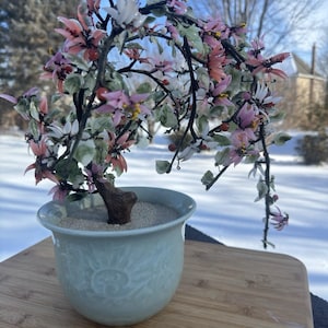 May include: A decorative bonsai tree with pink, white, and green glass flowers and leaves. The tree is planted in a light blue ceramic pot with a floral design. The pot sits on a wooden surface, with a snowy background.