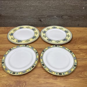 Op de afbeelding: Een set van vier ronde keramische borden met een wit midden en een decoratieve rand. De bovenste twee borden hebben een gele rand met blauwe bloemmotieven, terwijl de onderste twee borden een geel en groenblauw geometrisch ruitpatroon hebben.