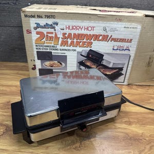 Op de afbeelding: Een zilveren en zwarte 2-in-1 sandwich- en pizzellemaker met de tekst "Hurry Hot" en "Model No. 795TC" op de doos. Het apparaat heeft een signaallampje en een zwarte handgreep. De doos is gemaakt in de VS.