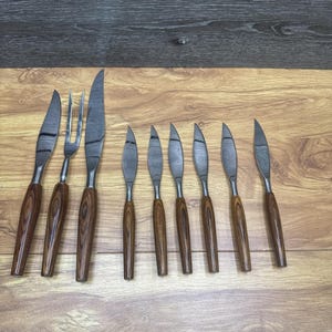 Könnte beinhalten: Ein Set aus Edelstahlmessern und einer Gabel mit Holzgriffen. Die Klingen haben ein strukturiertes Muster. Die Griffe sind dunkelbraun. Das Set enthält ein großes Messer, eine Gabel und mehrere kleinere Messer.