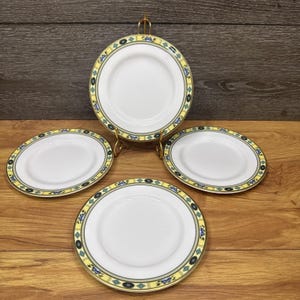 Op de afbeelding: Een set van vier witte keramische borden met een decoratieve rand met een gele band, blauwe bloemaccenten en geometrische ruitvormen in groenblauw en zwart. Eén bord staat rechtop op een gouden metalen standaard, drie liggen plat.