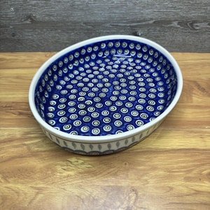 May include: An oval-shaped ceramic serving dish with a white rim and a deep blue interior. The interior is decorated with a repeating pattern of white and blue concentric circles. The exterior features a decorative border with a repeating pattern of gray and white leaf-like shapes.