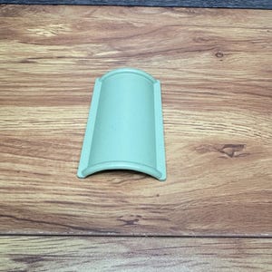 Puede incluir: Un objeto rectangular verde claro con la parte superior e inferior curvadas. El objeto está hecho de un material sólido y descansa sobre una superficie de madera. El fondo es de madera más oscura.