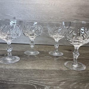 May include: Four clear crystal champagne coupe glasses. Each glass features a detailed, etched floral pattern on the bowl and stem. The glasses are displayed on a wooden surface, showcasing their elegant design.
