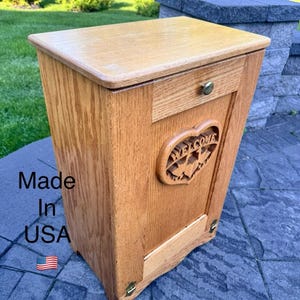 May include: A wooden trash can with a drawer and a carved heart design that says "WELCOME". The can is made of light brown wood and has brass hardware. The text "Made in USA" and an American flag are visible.