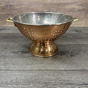 May include: A vintage copper and stainless steel colander with a pedestal base and two small handles. The colander has a perforated bowl and is designed for draining liquids from food. The copper has a warm, reddish-brown hue.