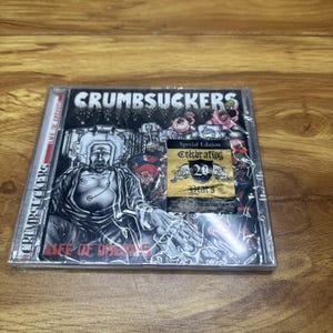 Op de afbeelding: Een Crumbsuckers Life of Dreams cd in een doorzichtig plastic doosje. De hoes bevat zwart-witte grafische kunst van een figuur met een mechanisch hoofddeksel, rode tekst Life of Dreams en een gouden sticker met de tekst Celebrating 20 Years.