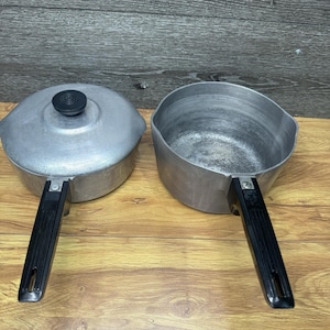 Puede incluir: Dos cacerolas vintage de aluminio con asas negras. Una cacerola tiene una tapa con un pomo negro. La otra cacerola está abierta. Ambas cacerolas están sobre una superficie de madera.