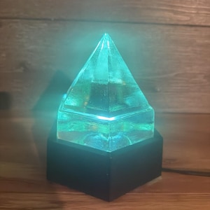 May include: A clear, pyramid-shaped object with a textured surface that emits a bright, glowing teal light. The pyramid is mounted on a solid, dark-colored hexagonal base, creating a contrast between the illuminated top and the dark support.