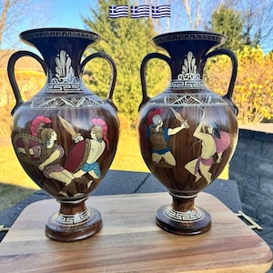 May include: Two brown ceramic vases with handles, featuring painted scenes of ancient Greek warriors. The vases have a dark brown rim and base, with gold and white decorative accents. The text "Made in Greece" is above the vases.