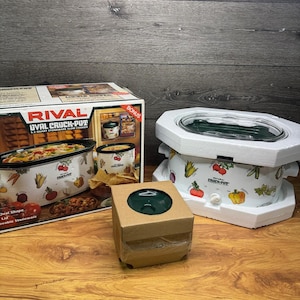 May include: A white Rival Crock-Pot slow cooker with a clear lid, and a cardboard box containing a green lid. The slow cooker has a decorative pattern of vegetables. The product is on a wooden surface.
