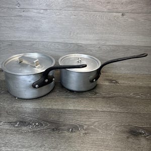 May include: Two aluminum saucepans with lids. The larger pan has a rectangular handle, while the smaller pan has a long, curved handle. Both pans have black handles and are sitting on a wooden surface. The pans are likely vintage kitchenware.