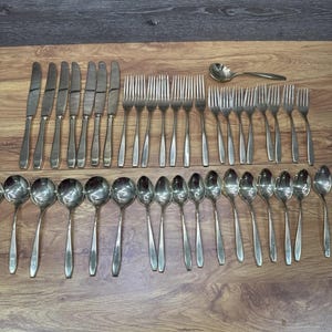 May include: A collection of stainless steel silverware, including knives, forks, and spoons, arranged on a wooden surface. The set includes various sizes of spoons, forks, and knives, ideal for a formal dining setting.