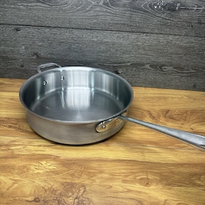 May include: Stainless steel frying pan with a long handle and a small loop handle on the opposite side. The pan has a shiny, metallic finish and is set against a wooden background.