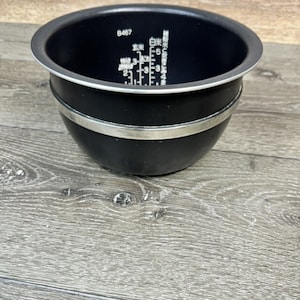 Może przedstawiać: Czarny garnek wewnętrzny do ryżowaru ze srebrną opaską i białym obramowaniem. Garnek ma oznaczenia pomiarowe w japońskich znakach. Garnek stoi na drewnianej powierzchni.