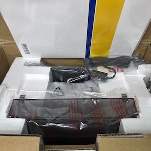 Puede incluir: Una caja de cartón que contiene un dispositivo electrónico negro envuelto en plástico transparente, piezas protectoras de poliestireno blanco y un cable de alimentación negro. Se ve un manual blanco con una franja azul y amarilla.