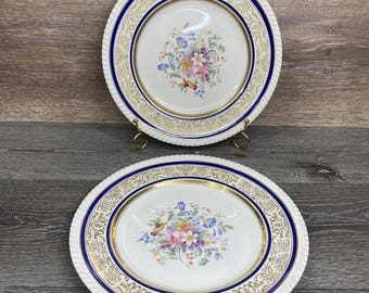 May include: Two white ceramic plates with a floral design. Each plate has a gold rim, a blue border, and a floral bouquet in the center. The plates are round and oval, and are displayed on a wooden surface.