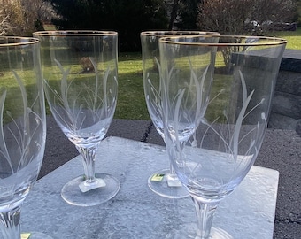 Può includere: Set di bicchieri da vino trasparenti con bordi dorati e disegni floreali incisi. I bicchieri hanno una forma classica con stelo e base ampia. Il design inciso presenta alte e sottili lame d'erba. I bicchieri sono esposti su una superficie grigia.