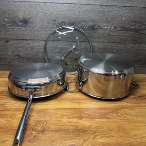 May include: Stainless steel cookware set featuring two saucepans and a glass lid. The saucepans have a copper-colored band around the top and a long handle on one. The glass lid has a silver handle. The set is displayed on a wooden surface.