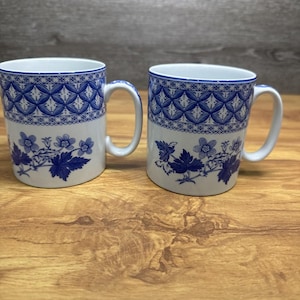 May include: Two white ceramic mugs with blue floral and geometric patterns. The mugs have a handle and a blue rim. The design includes blue flowers and leaves. The mugs are on a wooden surface.