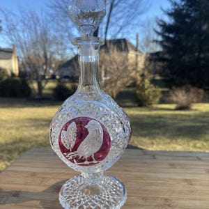 May include: A clear glass decanter with a diamond-cut pattern and a round base. The decanter features a bird and leaf design in a circular, maroon-coloured accent. The stopper is a clear, round, faceted ball.
