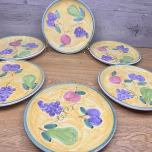 May include: A set of six ceramic plates with a yellow background and a hand-painted fruit design. The plates feature illustrations of purple grapes, green pears, and pink apples. Each plate has a blue rim.