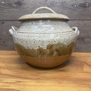 May include: A handmade ceramic lidded pot with a speckled, off-white glaze. The pot features a rounded shape with two side handles and a matching lid with a loop handle. The lower portion is a warm brown color.